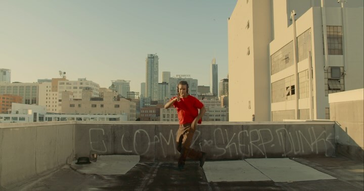 Adult male in a red shirt and brown pants is dancing on a rooftop with graffiti in an urban environment with buildings and skyscrapers.