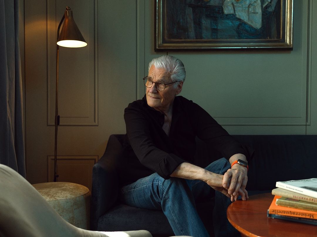 Elderly man wearing glasses, black shirt, and blue jeans seated on a dark sofa in a living room interior photographed by Yannick Wolff.