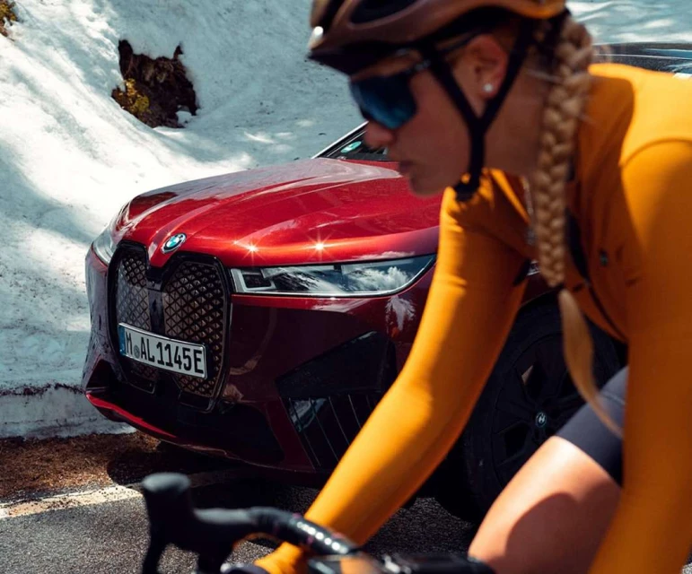Profile of a cyclist in a mustard-colored jersey, riding past a red BMW car on a paved road with a snow-covered background.