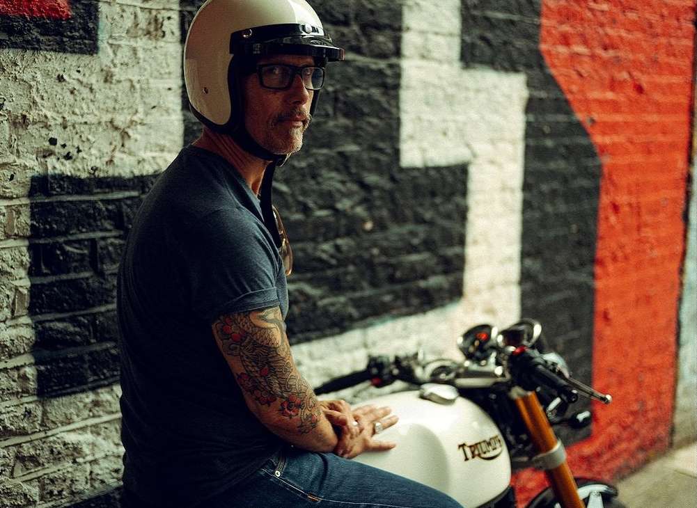 An adult man wearing a helmet and glasses sits on a white Triumph motorcycle in front of a brick wall painted with black, white and red.