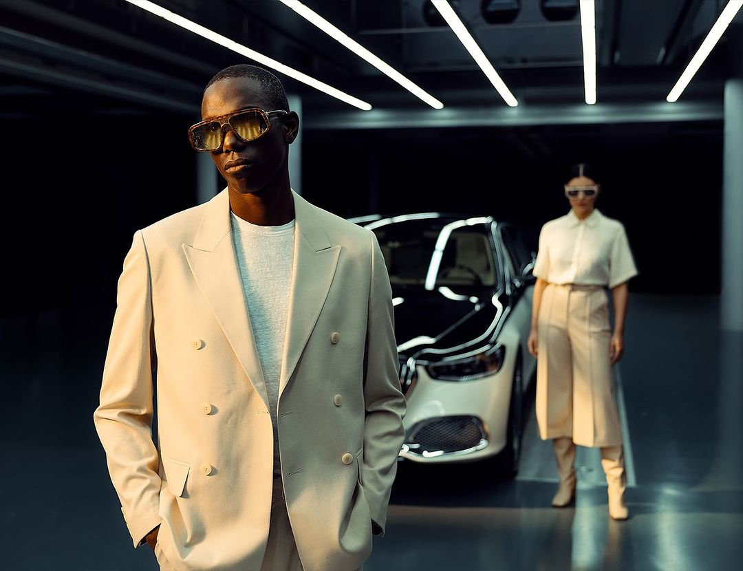 Two fashion models pose in a garage with a luxury car in the background, showcasing elegant beige and white outfits with stylish sunglasses.