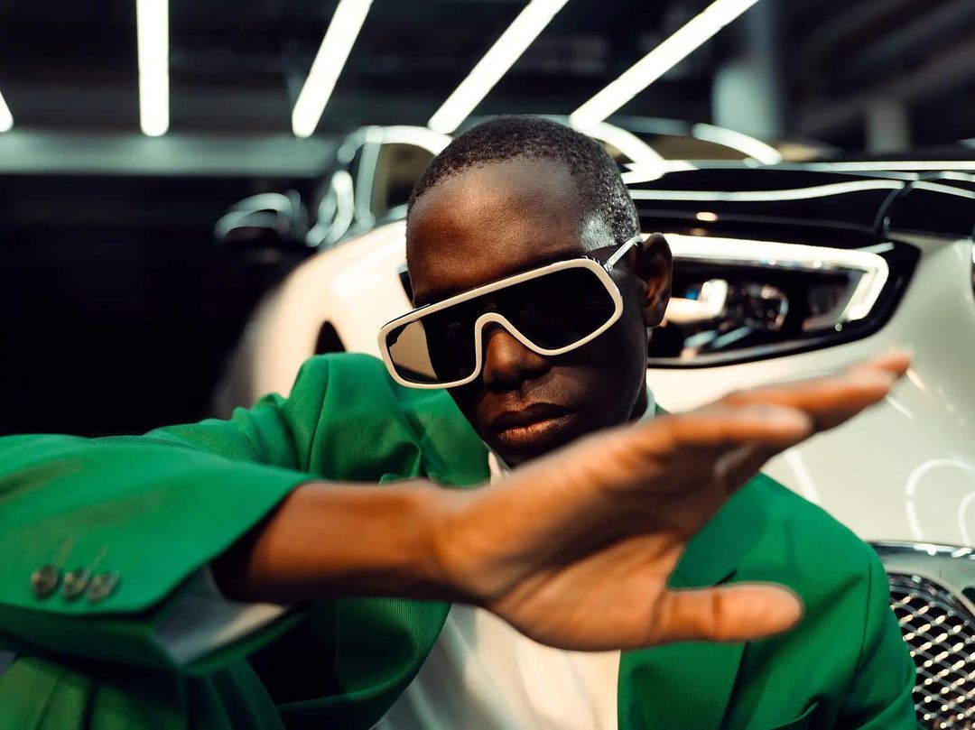 An adult male in a green suit and sunglasses extends his hand toward the viewer in front of a white car in an indoor setting.