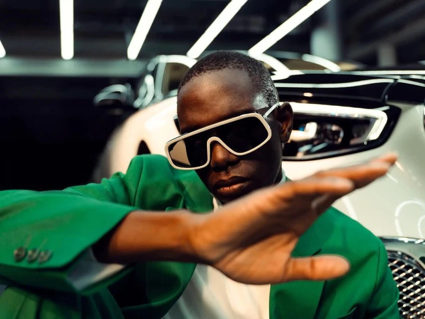 An adult male in a green suit and sunglasses extends his hand toward the viewer in front of a white car in an indoor setting.