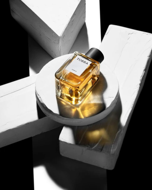 Product shot of a square glass perfume bottle with golden liquid on a white platform surrounded by abstract concrete blocks.