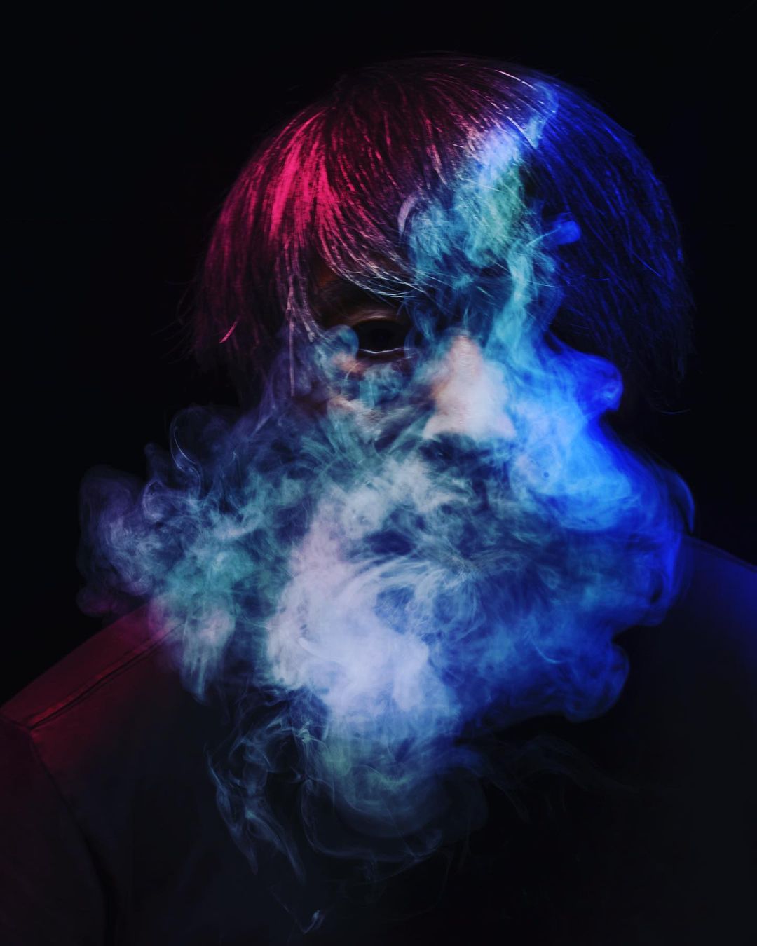 Portrait of a person with dark hair with pink and blue lighting effects and smoke or fog emanating from the face on a dark background.