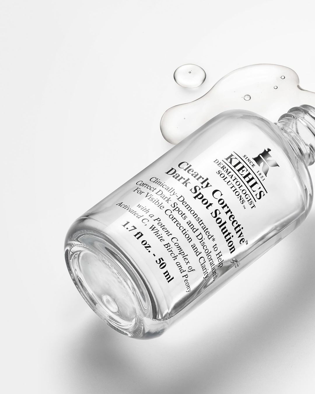 Cosmetic product shot: a Kiehl's Clearly Corrective Dark Spot Solution bottle on a white surface with clear liquid drops nearby.