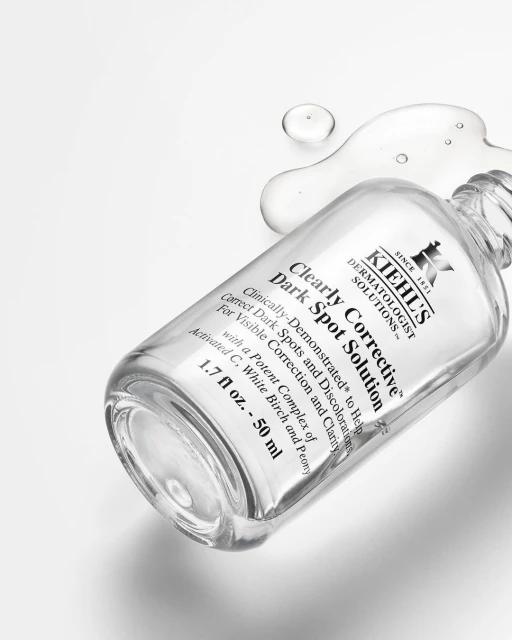 Cosmetic product shot: a Kiehl's Clearly Corrective Dark Spot Solution bottle on a white surface with clear liquid drops nearby.