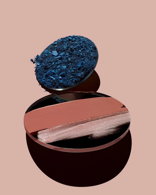 Beauty product still life: Blue eyeshadow crumbs and nude lipstick swatch arranged on reflective surfaces, set against a blush-colored background.