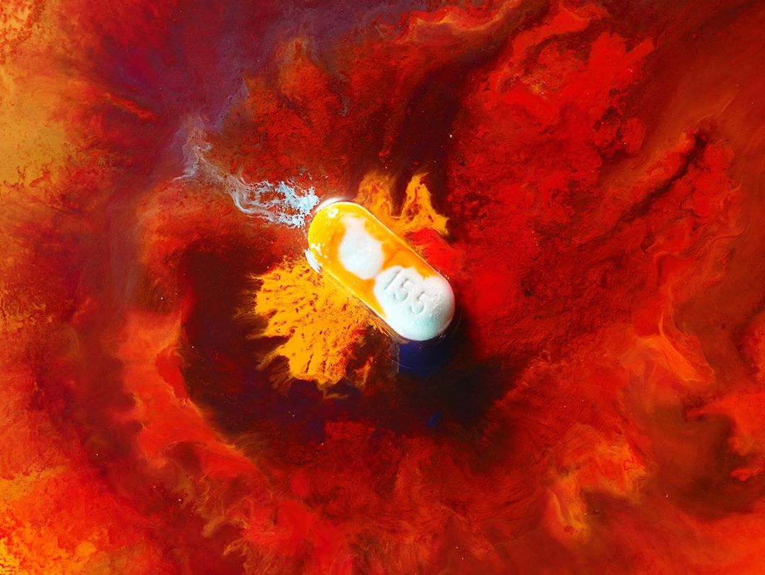 Capsule shaped medicine with imprinted numbers floats on vibrant abstract background of red, orange, and yellow pigment swirling together.