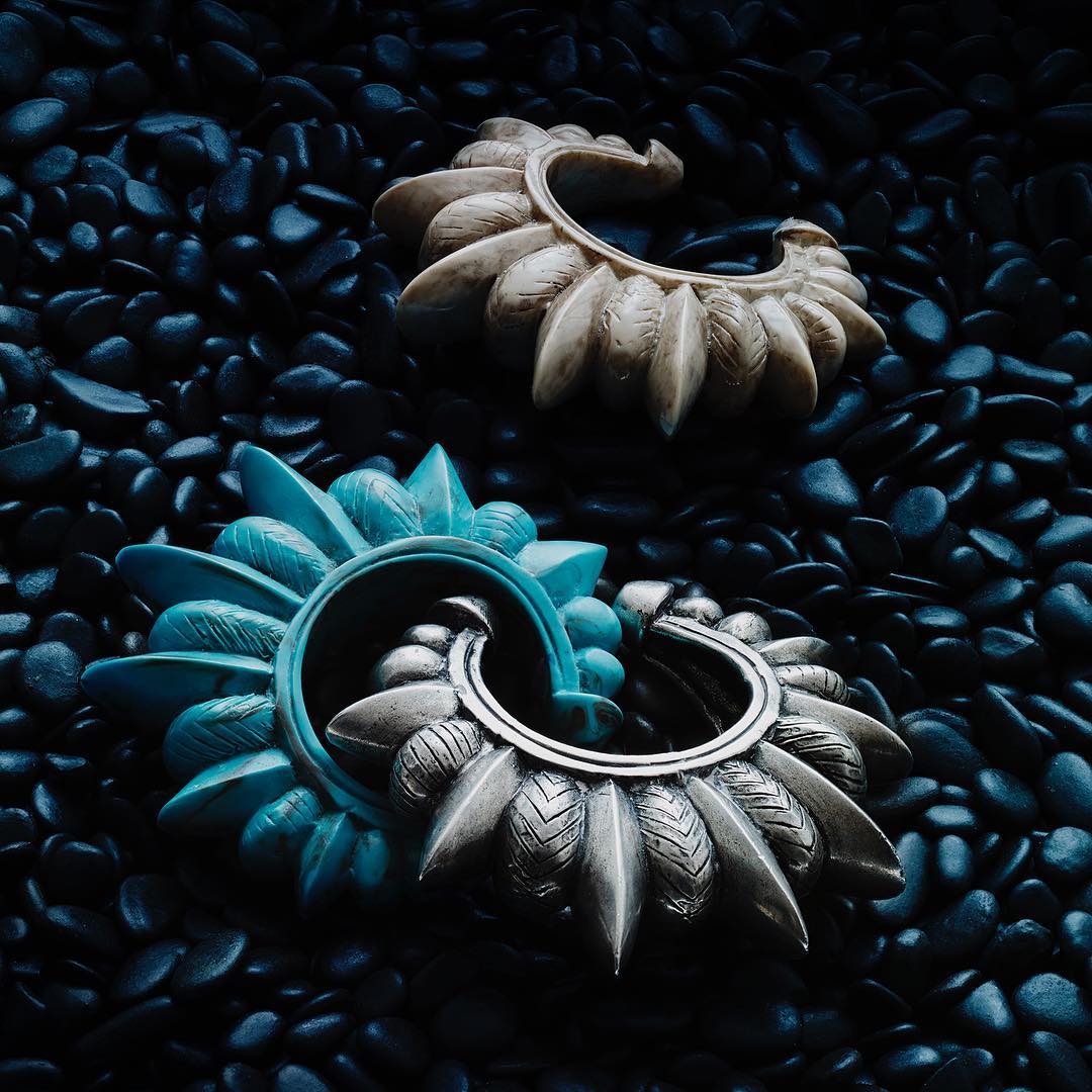 Two decorative semi-circular objects, one in teal and silver, the other cream, lie on a bed of small, polished black stones. 