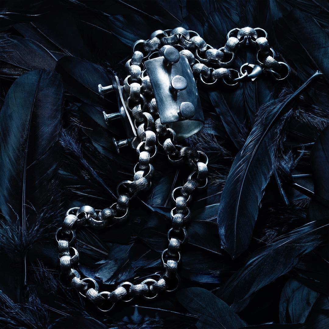 Elevated shot showcasing a silver chain necklace with a rectangular pendant against a backdrop of densely packed black feathers.
