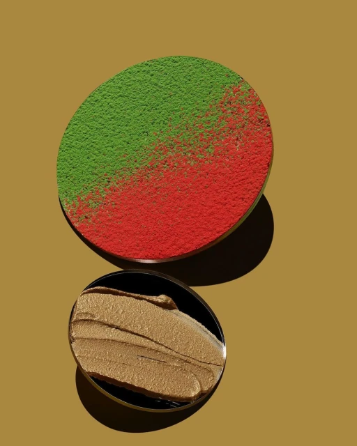 Overhead shot of a makeup palette with green and red powder, and a smear of gold cream make-up in black, round containers.