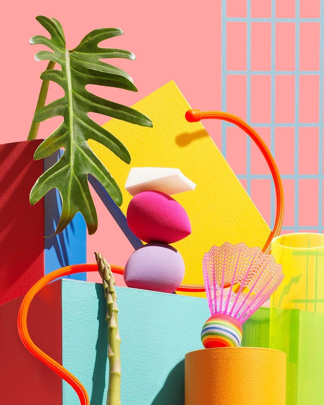 Beauty still life photography featuring makeup sponges, a tropical leaf, a badminton shuttlecock, and asparagus in a vibrant studio setup.