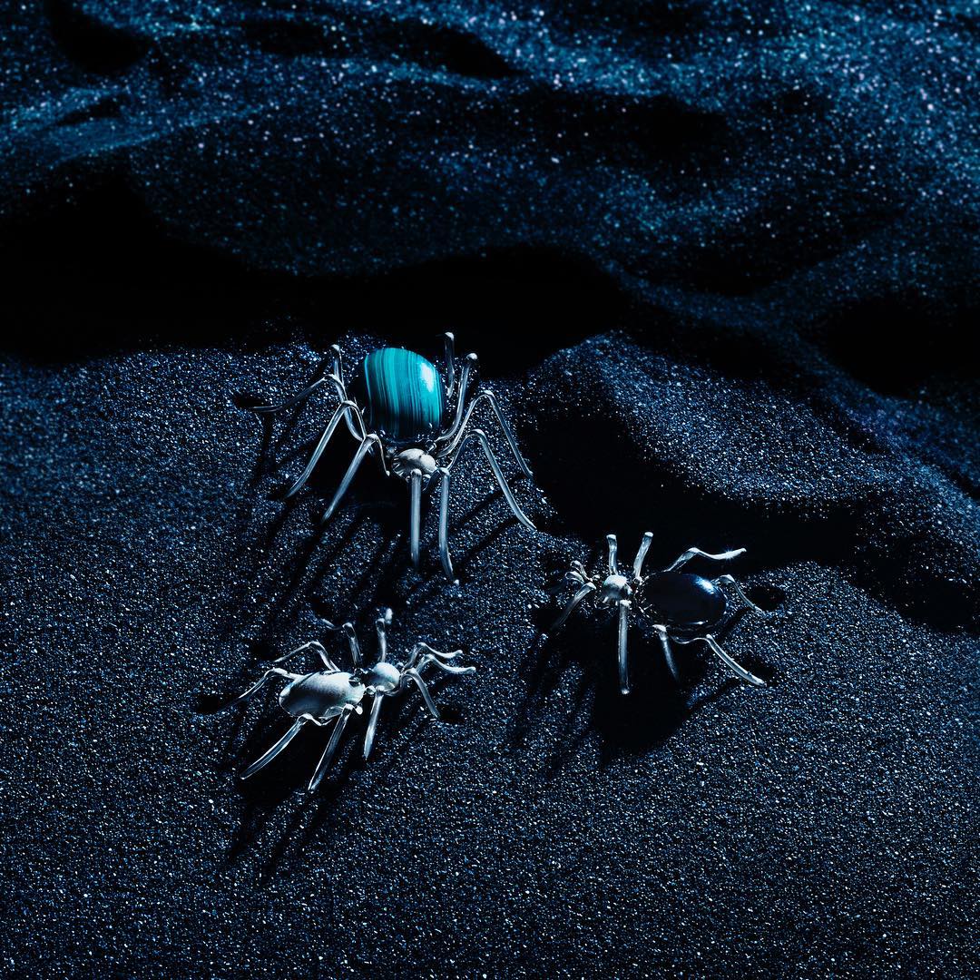 Three glass spider figures with colored accents stand on a surface of sparkling black sand, creating a dark and mysterious scene.