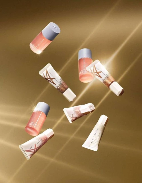 Cosmetic tubes and bottles from Fenty Beauty arranged in a visually appealing pattern against a gold-colored background with light rays.