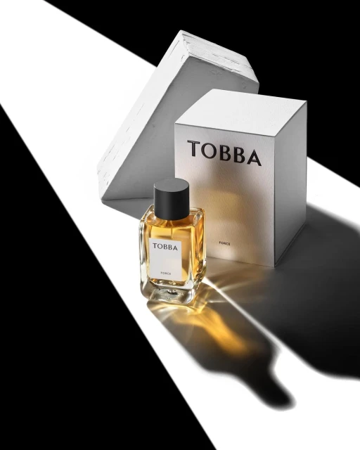 Studio shot of a clear glass bottle of amber perfume, placed near its white box, and abstract blocks with striking black-and-white lighting.