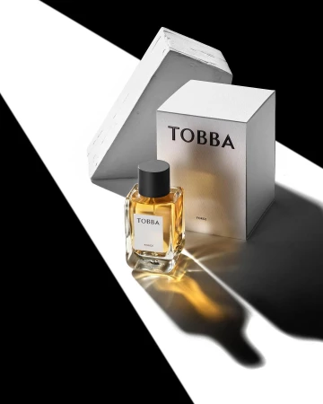 Studio shot of a clear glass bottle of amber perfume, placed near its white box, and abstract blocks with striking black-and-white lighting.