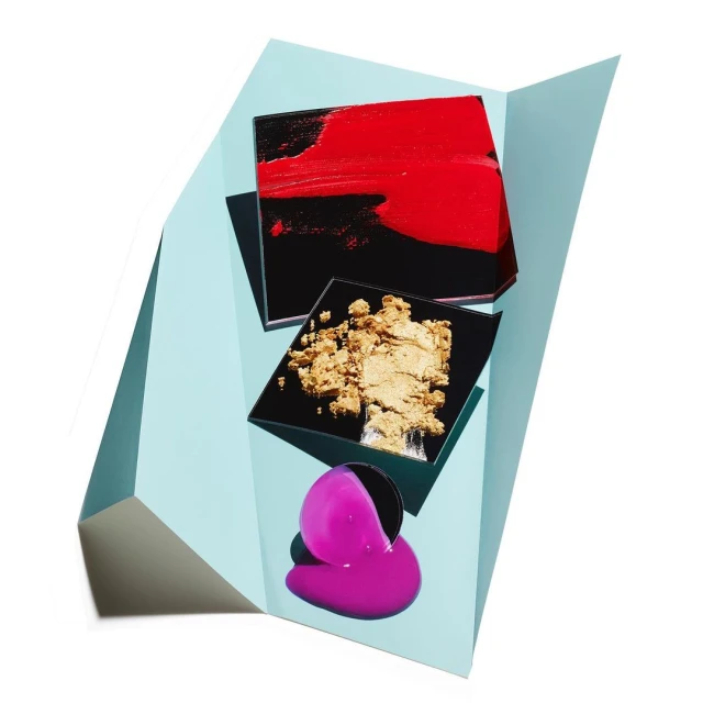 Cosmetic products including eye shadow, lip gloss, and lipstick displayed on a light blue geometric stage against a white backdrop.
