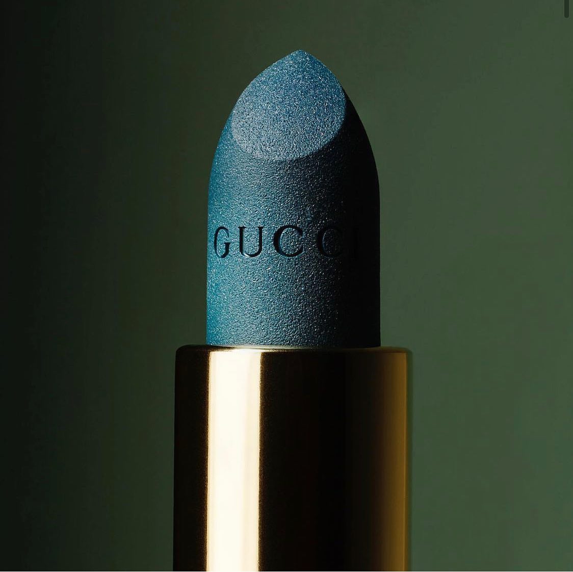 Close-up studio shot of a Gucci lipstick with a teal bullet and gold tube, presented against a soft green background.
