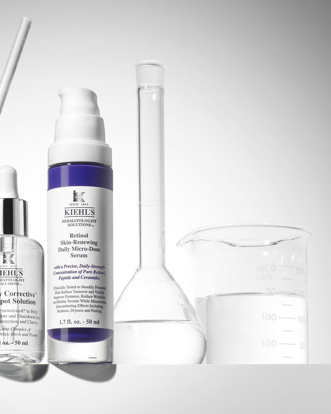 Kiehl's skincare products displayed in a laboratory-like setting, including retinol serum and spot solution bottles next to beakers.