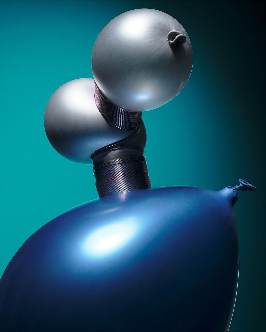 Sculptural arrangement of blue and silver balloons, resembling a futuristic form, set against a graduated teal backdrop. Smooth textures.