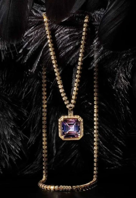 Gold necklace with a large square amethyst pendant on a chain. Jewelry is resting against a background of black feathers, high-key lighting.