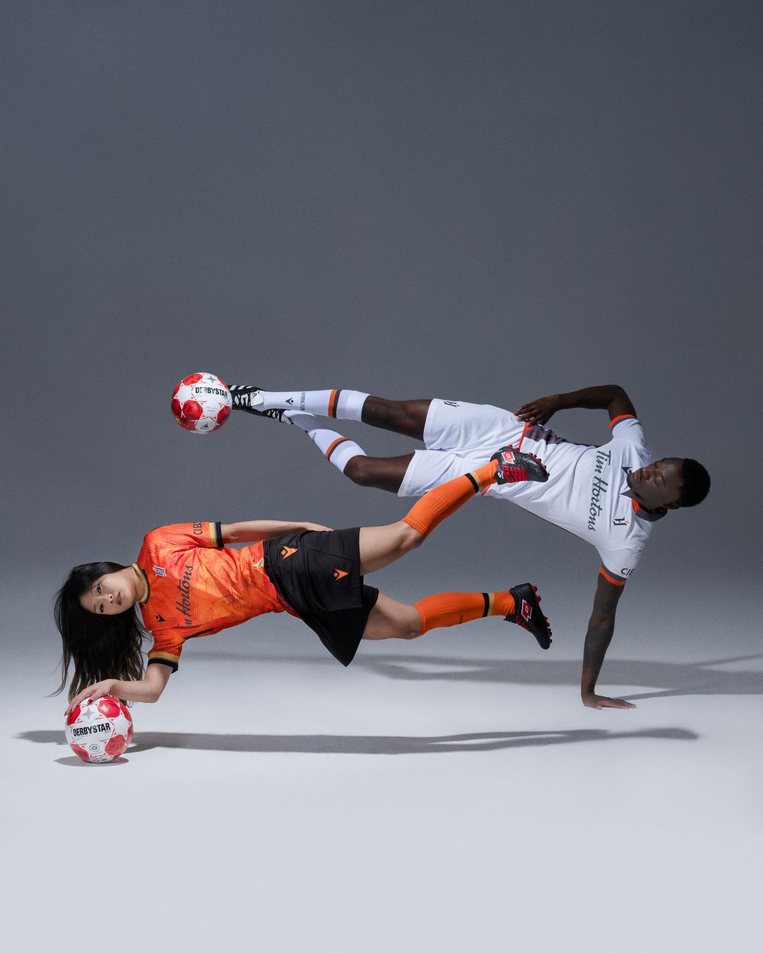 Two soccer players, one Asian female and one Black male, in mid-air pose with soccer balls in a studio with grey background.