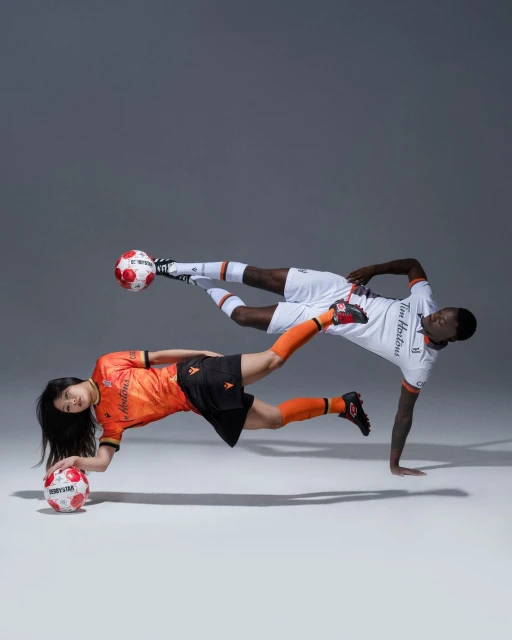 Two soccer players, one Asian female and one Black male, in mid-air pose with soccer balls in a studio with grey background.