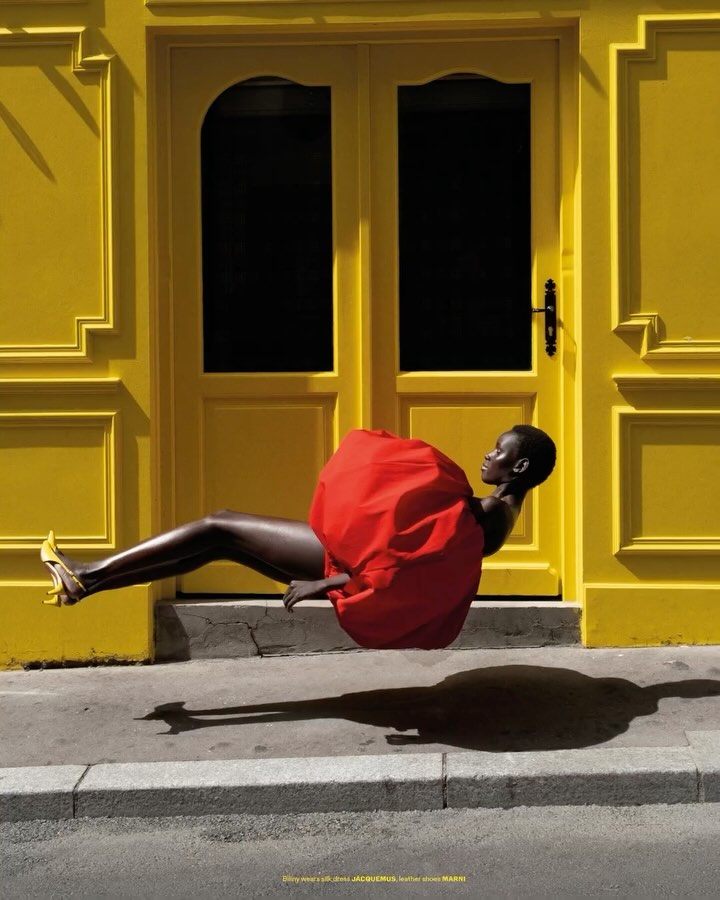 An elegant Black woman in a red dress and yellow high heels levitates in front of a yellow door and grey pavement casting a shadow.