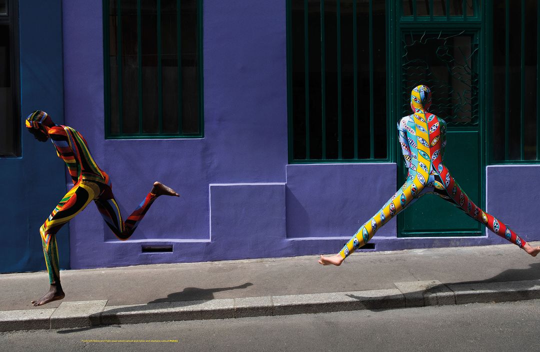 Two models wearing colorful body suits appear to be running on a city street in front of a purple building with a green door and window.