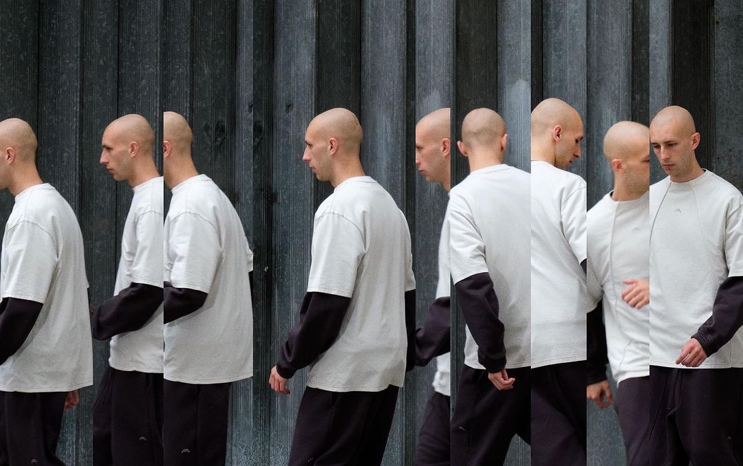 Multiple exposures of a shaved-head man wearing a white shirt with black long sleeves, walking against a textured gray wall.