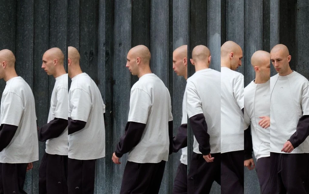 Multiple exposures of a shaved-head man wearing a white shirt with black long sleeves, walking against a textured gray wall.