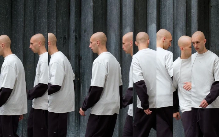 Multiple exposures of a shaved-head man wearing a white shirt with black long sleeves, walking against a textured gray wall.