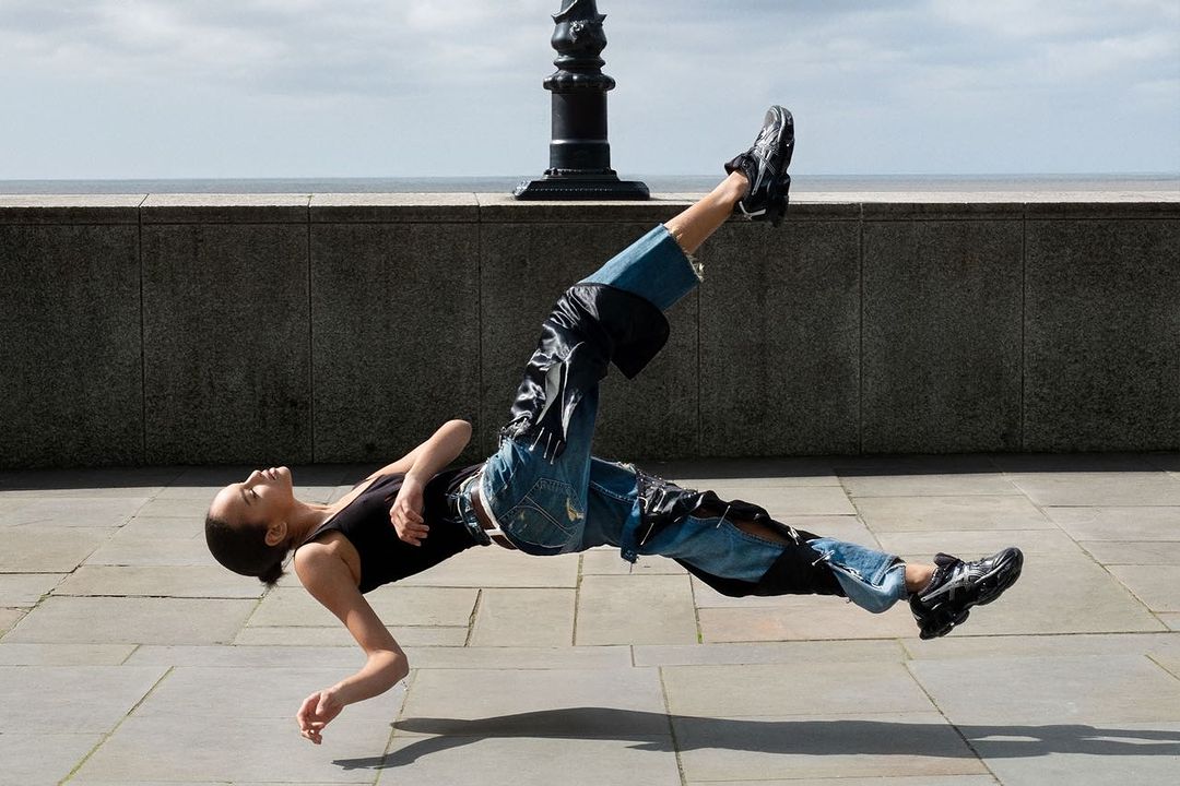An adult female levitates in an urban outdoor setting, wearing a black top, jeans, and sneakers, with a wall, ocean, and sky in the background.