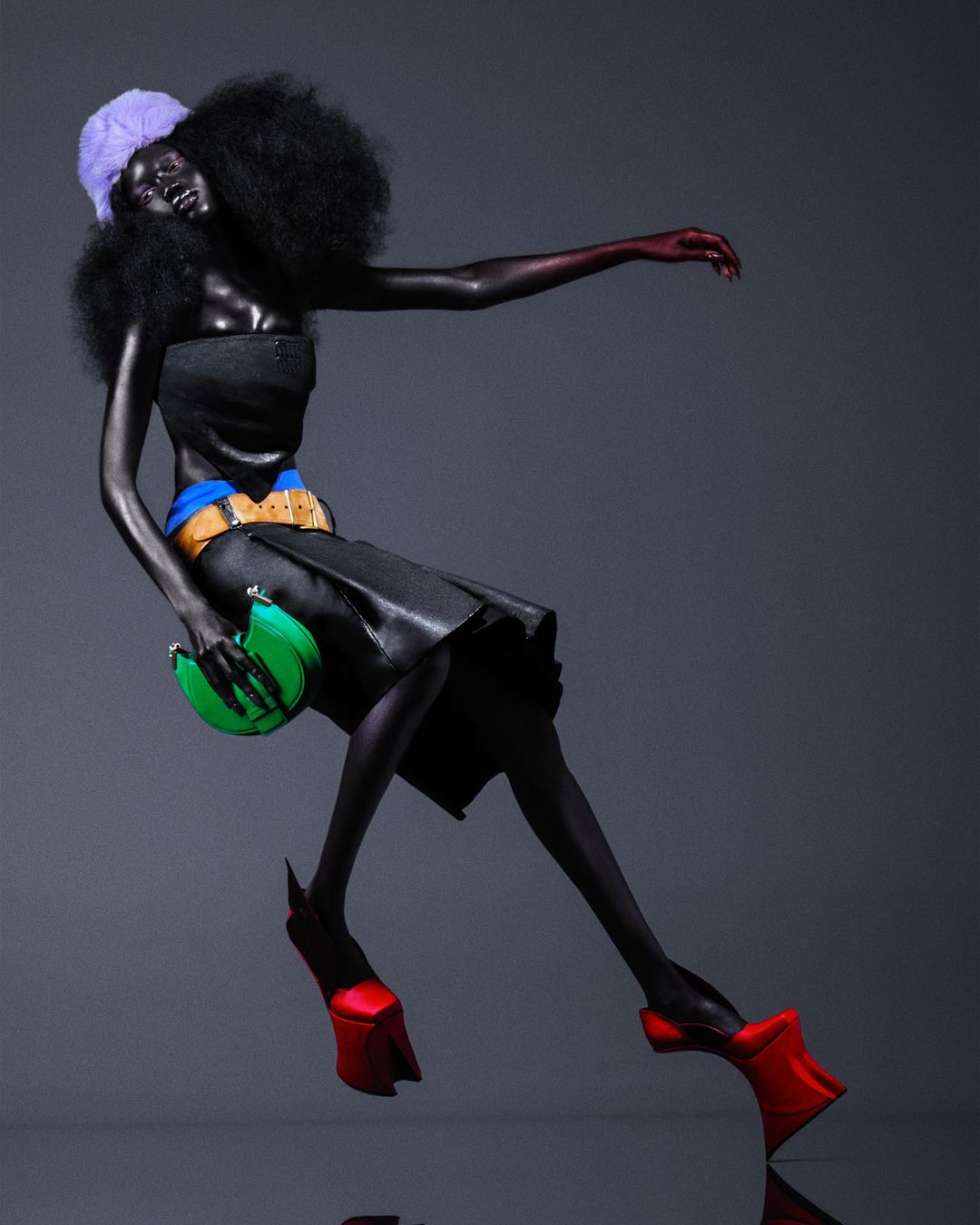 Fashionable adult female in black dress, red shoes, and purple hat posing in a studio. She is holding a green purse.