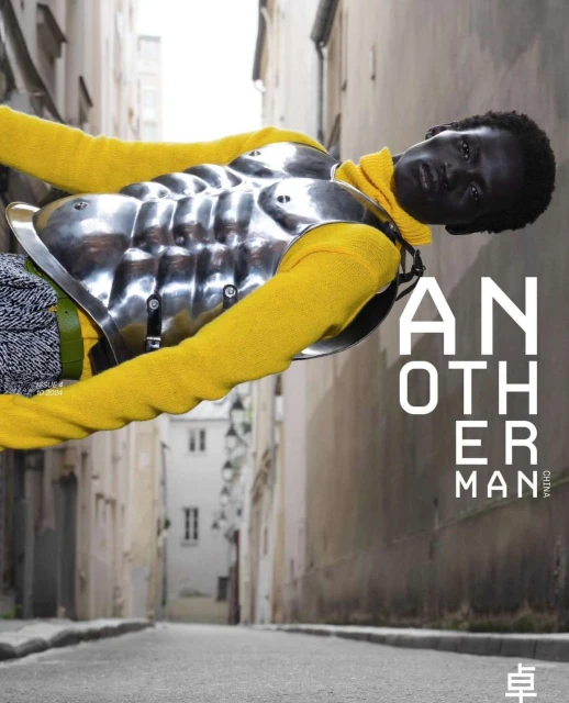 Fashion editorial shot of a Black model wearing yellow turtleneck and metal armor in a European city street, leaning on building with text.