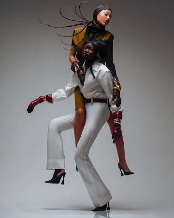 Two fashion models posing together against a grey studio backdrop, one piggybacking on the other in dynamic fashion photography.