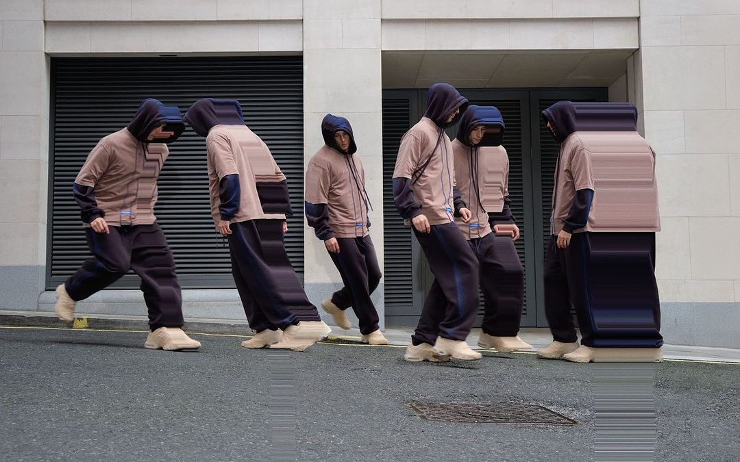 Multiple figures in hooded sweatshirts and sweatpants are standing on a city street with a grey brick building in the background.