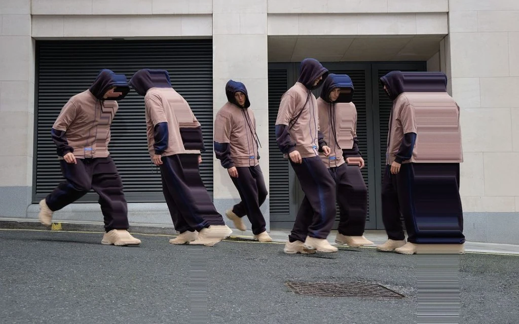 Multiple figures in hooded sweatshirts and sweatpants are standing on a city street with a grey brick building in the background.