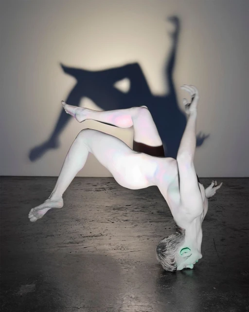 Artistic studio shot of a person covered in white body paint posing upside down with their shadow projected on a white wall.