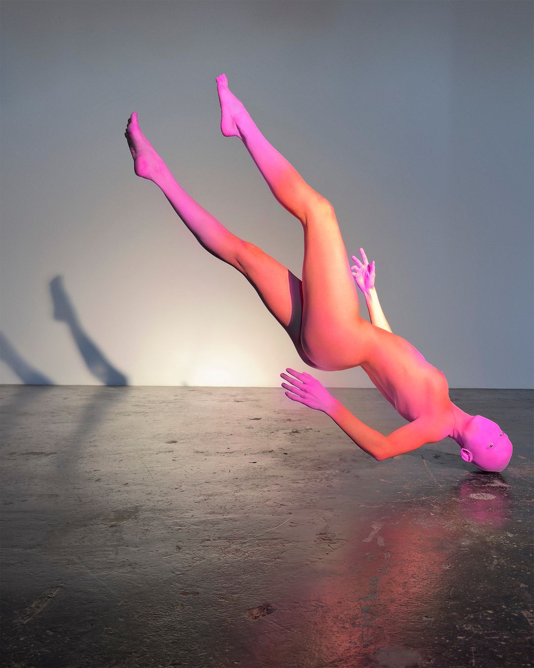Full body shot of a pink female figure lying on her back, with pink-ombre legs up, on a dark concrete floor with a light grey background.