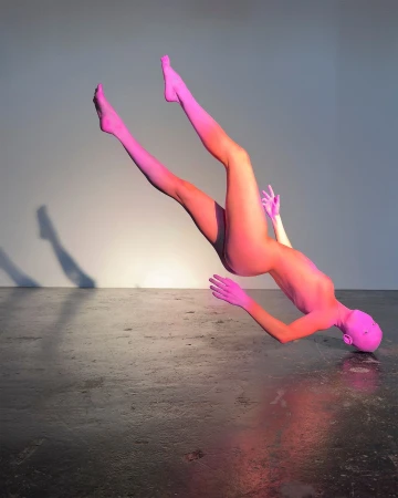 Full body shot of a pink female figure lying on her back, with pink-ombre legs up, on a dark concrete floor with a light grey background.