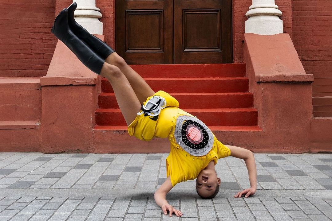 An androgynous adult doing a handstand in black boots and yellow garment with a decorative circular element, on an urban street.