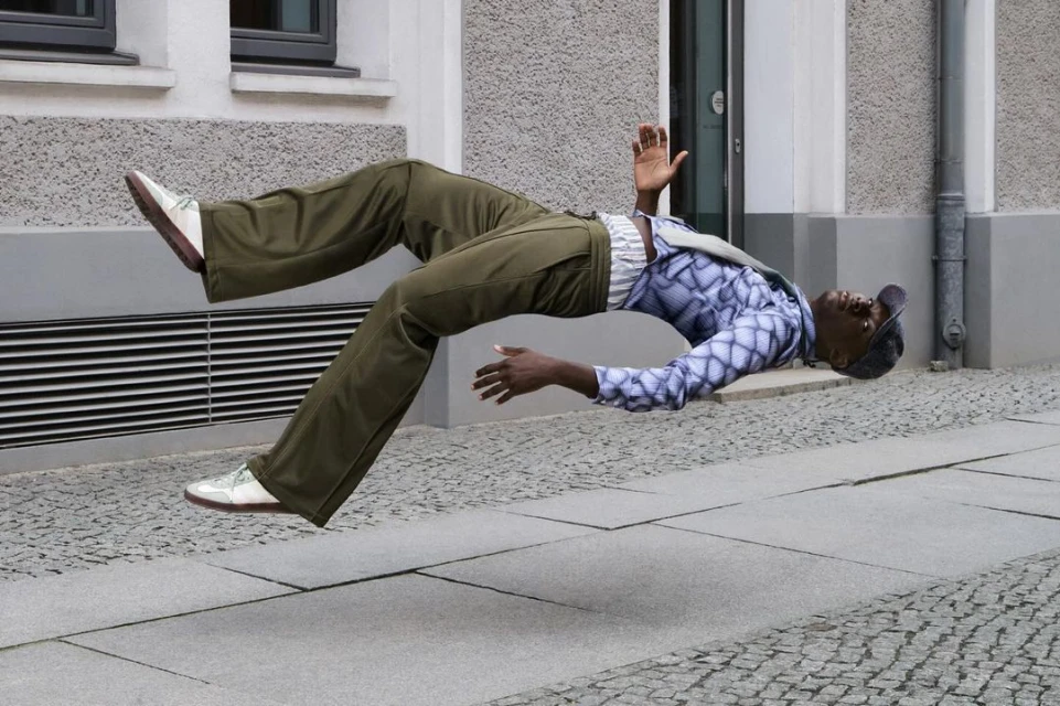 Young Black man floats horizontally in mid-air on a city sidewalk, wearing green pants, patterned shirt, and a gray cap. Building in background.