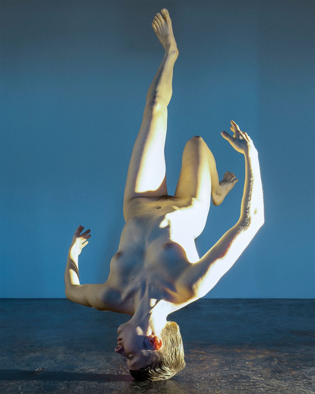 Nude adult leaning on head with feet up, facing forward against a split light blue and dark grey studio background in a portrait framing.