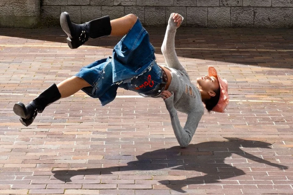 Woman in grey sweater and denim shorts performing a handstand in a black boots and a hat, shadow on the brick ground.