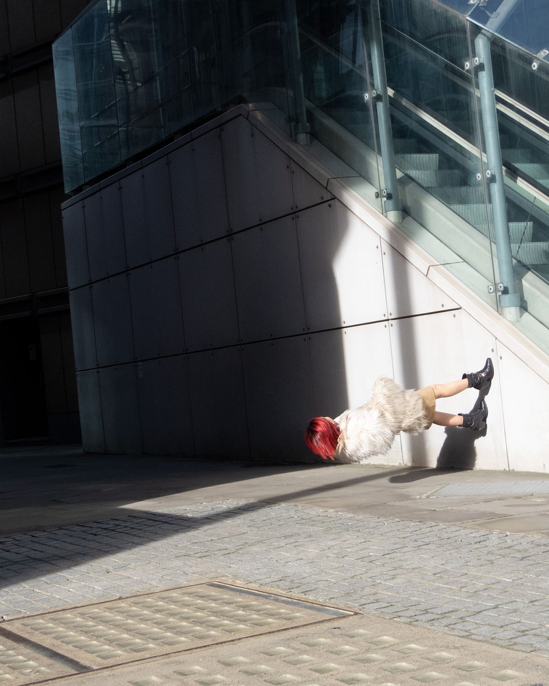 View of adult female lying on her side near building wall with glass stairway, wearing black boots and fur coat in outdoor urban setting.