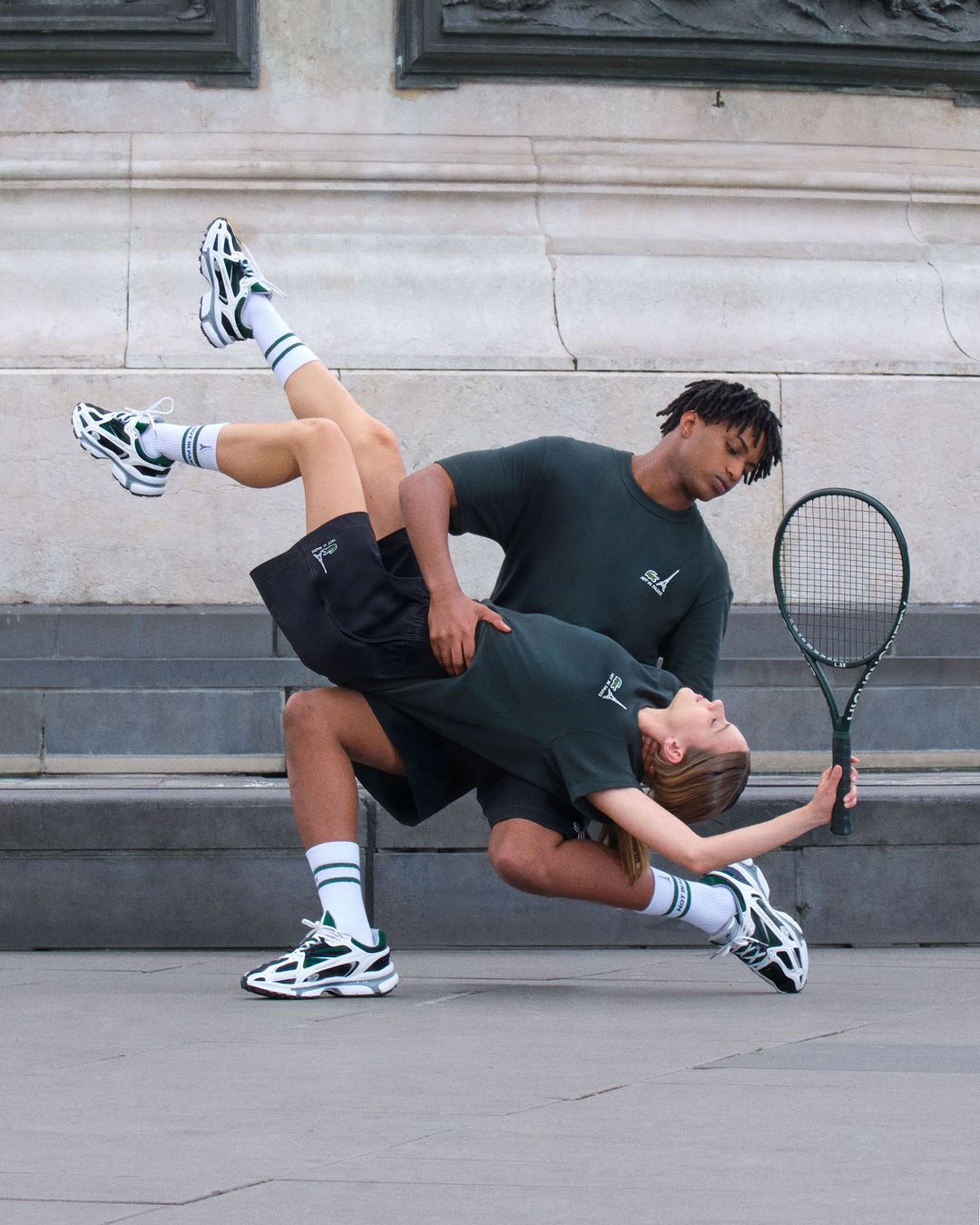 Two people in matching athletic apparel strike a dynamic pose with a tennis racket against a monumental stone wall outdoors.