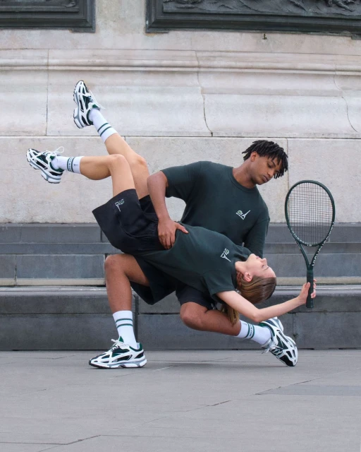 Two people in matching athletic apparel strike a dynamic pose with a tennis racket against a monumental stone wall outdoors.