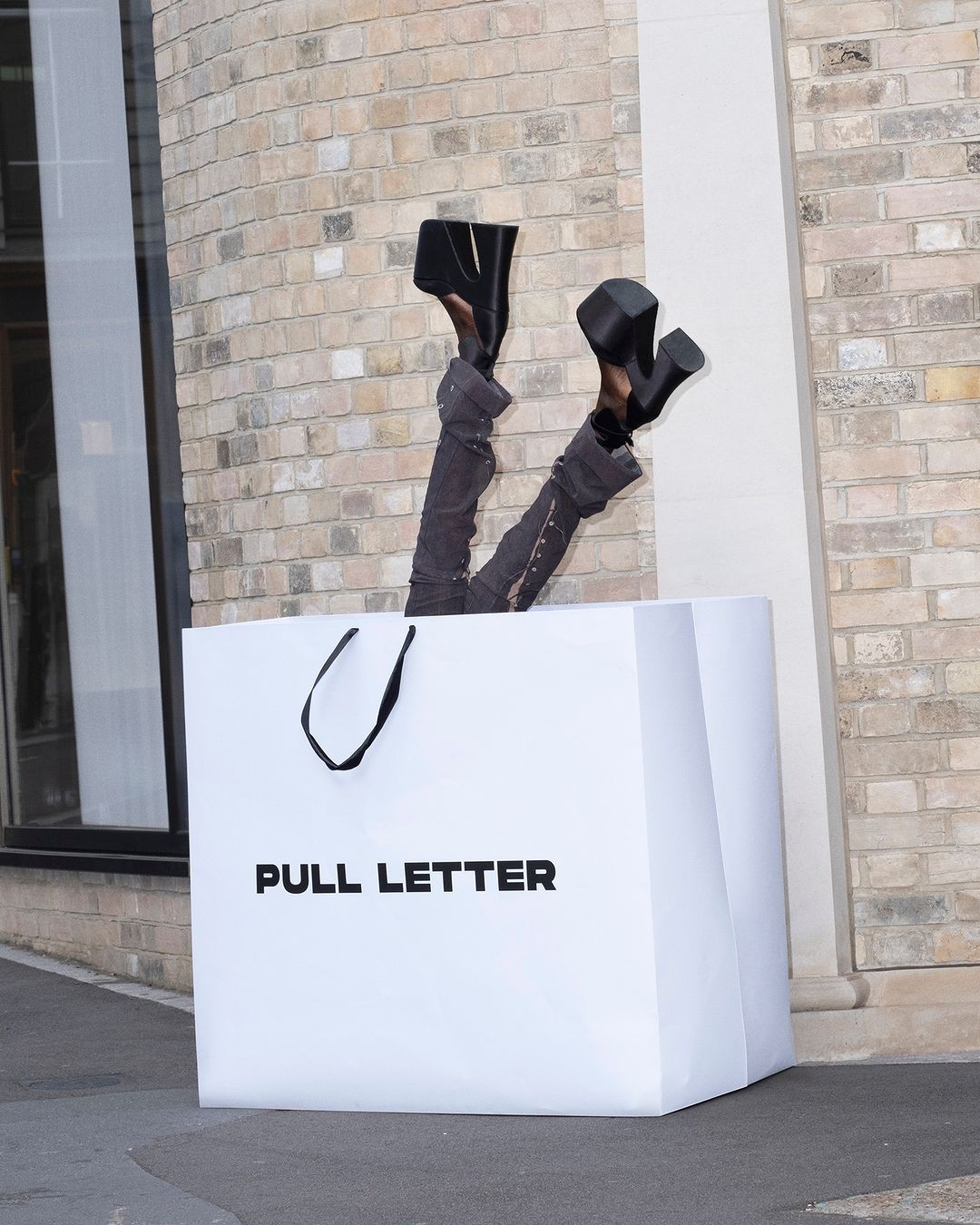 Legs in dark trousers and platform shoes sticking out of a large white shopping bag reading Pull Letter